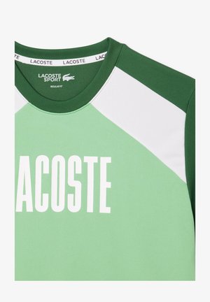 Camiseta deportiva verde con cuello redondo, paneles de hombros en blanco contrastante y gran texto blanco "LACOSTE". Diseño de ajuste regular.