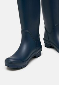 Tengerészkék, sima felületű gumibakancsok, masszív talppal, alacsony sarokkal és diszkrét varrásokkal. Nedves időjárási körülményekhez ideális.