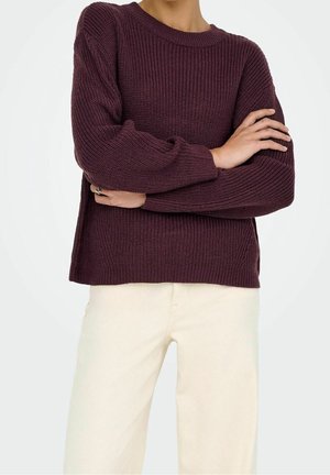 Personne portant un pull en laine violet foncé à texture côtelée et un pantalon crème, bras croisés à la taille.