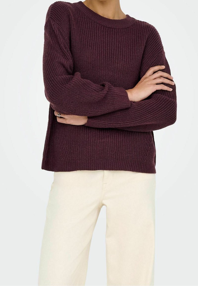 Personne portant un pull en laine violet foncé à texture côtelée et un pantalon crème, bras croisés à la taille.