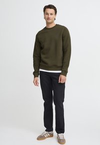 Olijfgroen sweatshirt met een ronde halslijn en ribgebreide manchetten, gecombineerd met zwarte rechte broek en grijze sneakers.