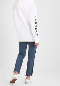 Personne portant un hoodie blanc avec logo sur la manche, un jean bicolore bleu avec des ourlets effilochés, et des baskets blanches en train de s'éloigner.