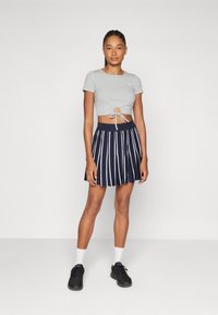 T-shirt grise coupée avec un nœud sur le devant et un découpage rond, associée à une jupe plissée bleu marine avec des rayures blanches. Baskets noires et chaussettes blanches.