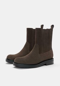 Bocanci din suede maro, cu panouri elastice pe lateral, vârf rotund neted și talpă din cauciuc negru. Prezintă două bucle în partea de sus pentru a fi ușor de purtat.