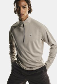 Šviesiai rudos ilgos rankovės sportinė palaidinė su ketvirčio užtrauktuku priekyje, juodas akcentinis užtrauktukas ir logotipas kairėje krūtinės pusėje; lygi tekstūra, prigludęs dizainas.