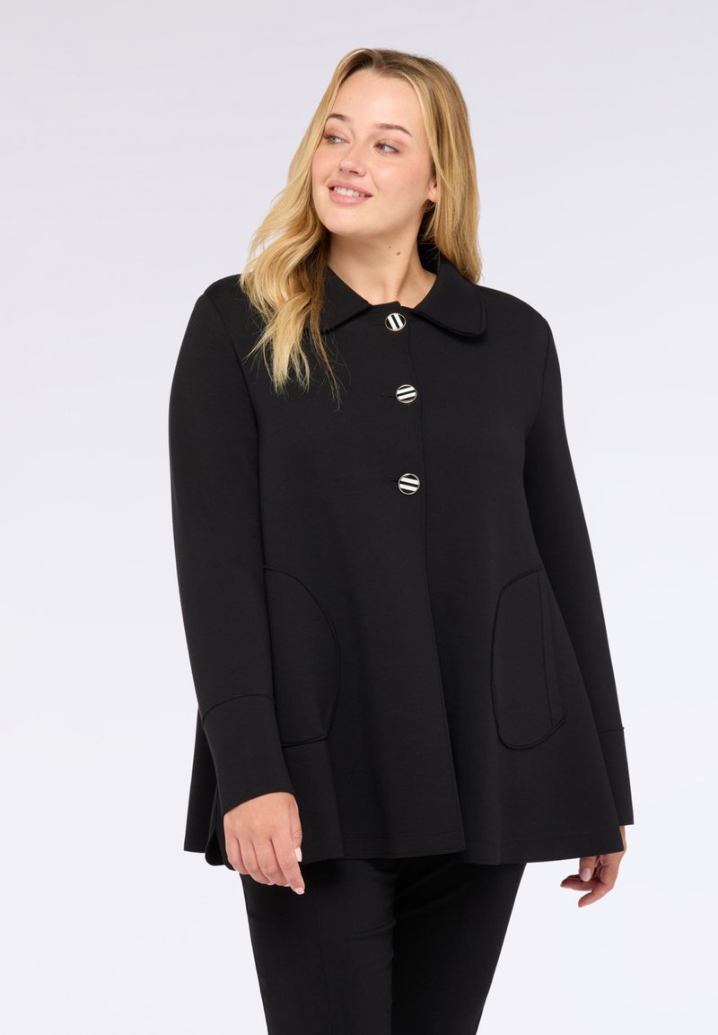 Veste noire en matériau doux, avec un col arrondi, quatre boutons ronds et des poches latérales. La coupe est ajustée et légèrement ample.