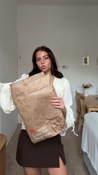 Brązowa torba papierowa z napisem "Kochaj mnie. Noś mnie." w białym i czerwonym kolorze, trzymana przez osobę w białej bluzce z koronkowymi detalami i czarnej spódnicy.