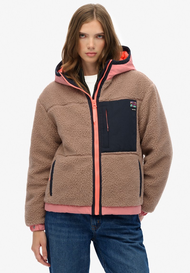 Bruine fleecejas met marineblauwe accenten, heldere koraalkleurige ritssluiting en capuchon. Voorzien van een voorzak, gestructureerde stof en elastische manchetten.