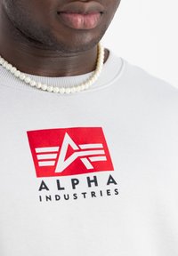 Felpa bianca in cotone con un logo rosso e nero ben visibile con la scritta "ALPHA INDUSTRIES", e una collana di perle attorno al collo.