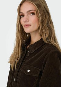 Jauna sieviete ar gariem viļņainiem blondiem matiem, valkājot tumši brūnu kordurojas jaku ar sudraba pogām, maigi skatās kamerā.