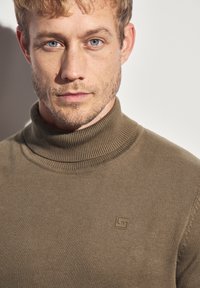 Maglione girocollo marrone con una texture a coste, caratterizzato da una piccola etichetta del logo sul davanti. Dettaglio ravvicinato del tessuto e del colletto.