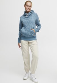 Sudadera azul con cuello de cowl, tres botones y un patrón floral; combinada con pantalones jogger beige y zapatillas blancas.