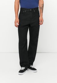 Jeans de mezclilla negra con un corte relajado, cinco bolsillos, cierre de botón de metal y diseño liso, combinados con zapatillas negras.