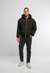 Felpa nera con zip, dotata di un cappuccio ampio, tasche frontali e polsini a costine, abbinata a jeans scuri e sneakers chiare.