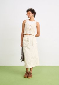 Kremowa kamizelka na szydełku z wiązaniem z przodu, noszona na białym topie na ramiączkach, zestawiona z beżową maxi spódnicą i brązowymi zamszowymi sneakersami. Zielona podłoga.