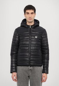 Chaqueta de plumas negra con capucha, con textura acolchada, un cierre frontal y un parche con el logo en el lado izquierdo del pecho. Llevada con jeans grises.
