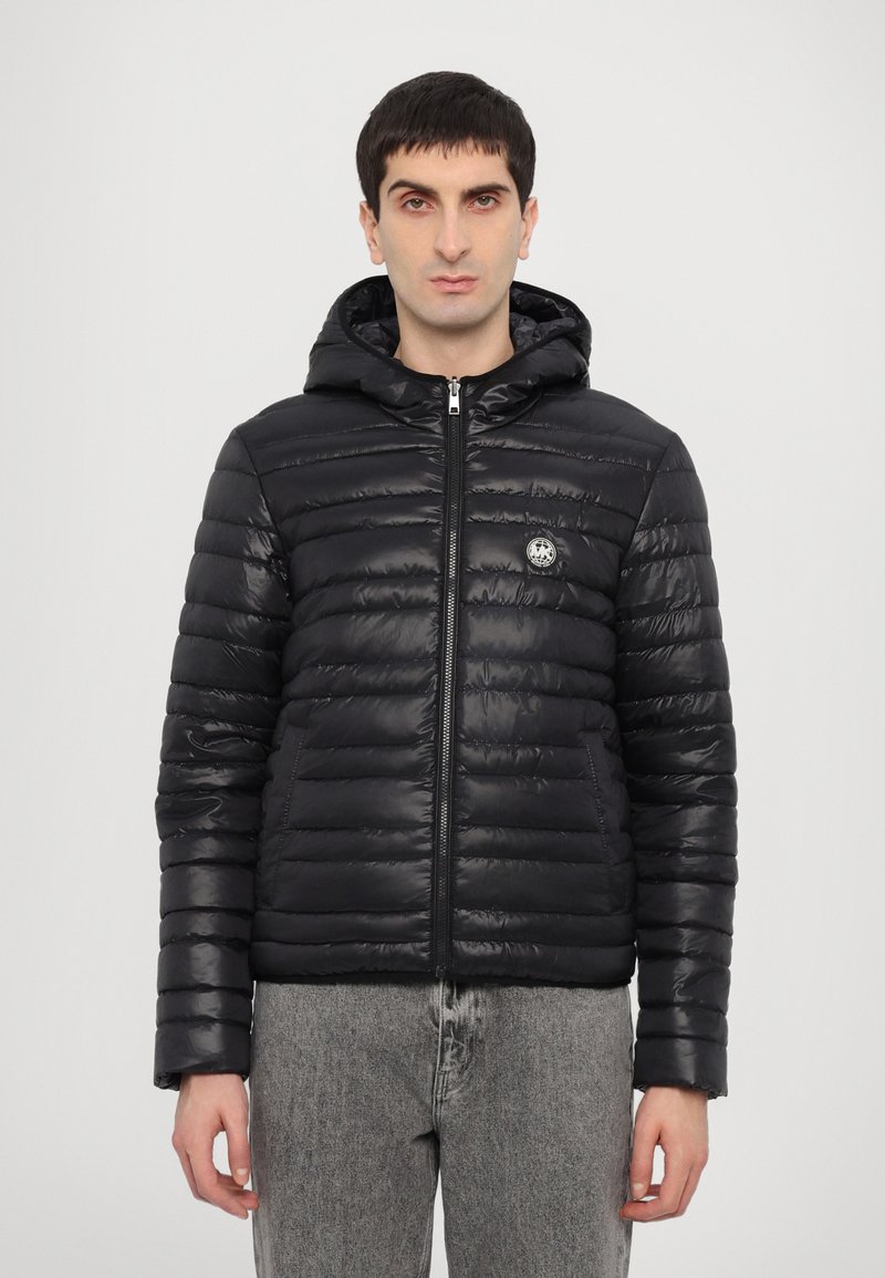 Chaqueta de plumas negra con capucha, con textura acolchada, un cierre frontal y un parche con el logo en el lado izquierdo del pecho. Llevada con jeans grises.