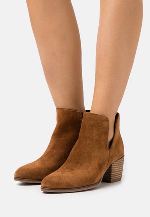Bruine suède enkellaarsjes met gestapelde blokhakken, gedragen op blote benen tegen een effen lichte achtergrond.