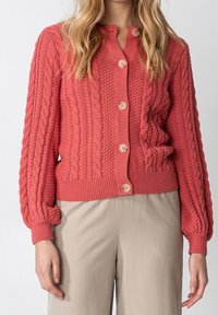 Koralfärgad stickad cardigan med kabelmönster, rund hals, långa ärmar och träknappar. Har ribbade manschetter och nederkant.
