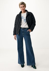 Schwarze Daunenjacke mit gestepptem Muster, weißes Grafik-T-Shirt, weit geschnittene blaue Jeans und grüne Absatz-Stiefeletten.