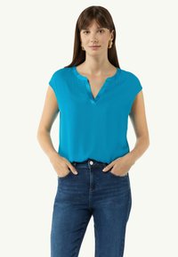 Türkise Kurzarmbluse mit V-Ausschnitt, aus glattem Stoff. Kombiniert mit dunkelblauen Jeans, Hände in die Hüften gestützt. Keine auffälligen Muster.