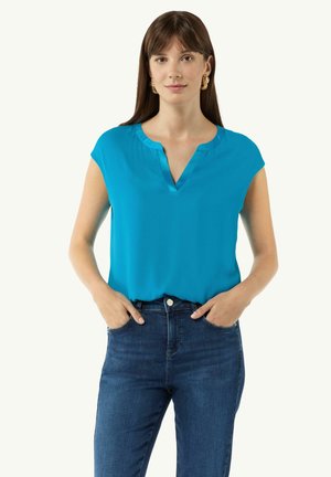 Turquoise short-sleeve blouse with a V-neckline, made of smooth fabric. Paired with dark blue jeans, hands on hips. No prominent patterns.