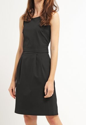 Ærmeløs sort knælængde kjole med tilpasset talje og lette læg, båret af en person med langt brunt hår mod en ensfarvet baggrund.