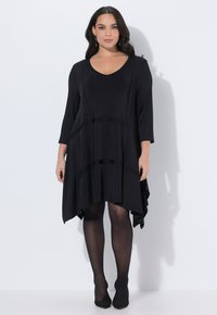 Nainen pukeutuneena mustaan, pitkähihaiseen, epäsymmetriseen helmainen mekkoon, jonka kanssa on päällä läpikuultavat mustat sukkahousut ja mustat teräväkärkiset kengät, seisomassa valkoista seinää vasten.