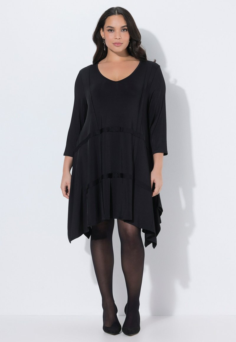 Nainen pukeutuneena mustaan, pitkähihaiseen, epäsymmetriseen helmainen mekkoon, jonka kanssa on päällä läpikuultavat mustat sukkahousut ja mustat teräväkärkiset kengät, seisomassa valkoista seinää vasten.