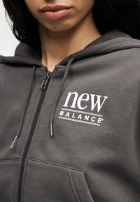 Κοντινό πλάνο ατόμου που φορά φούτερ με φερμουάρ σε γκρι απόχρωση, με λευκό λογότυπο "new balance" στο αριστερό μέρος του στήθους και κορδόνια να κρέμονται.