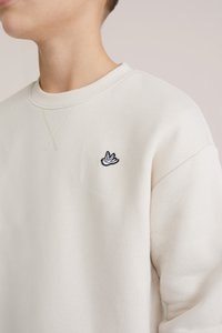 Bluză sport de culoare bej deschis, realizată dintr-un material moale, având un guler rotund și un mic logo de pasăre brodată negru pe partea stângă a pieptului.