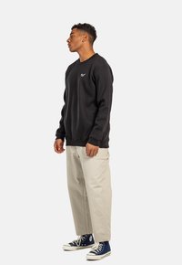 Schwarzer Sweatshirt mit gerippten Bündchen, kombiniert mit hellbeigen Hosen und blauen High-Top-Sneakern. Verfügt über ein kleines Logo-Detail vorne.