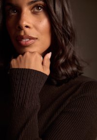 Un suéter marrón oscuro con textura acanalada y patrón texturizado, mangas largas que cubren parcialmente una mano con un anillo plateado, sobre un fondo oscuro.