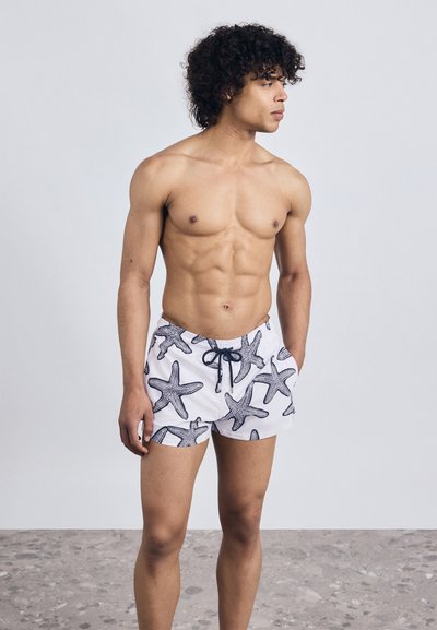 Homme torse nu avec des cheveux bouclés portant un short de bain blanc à motifs d'étoiles de mer noires, se tenant sur un sol gris moucheté devant un fond uni.