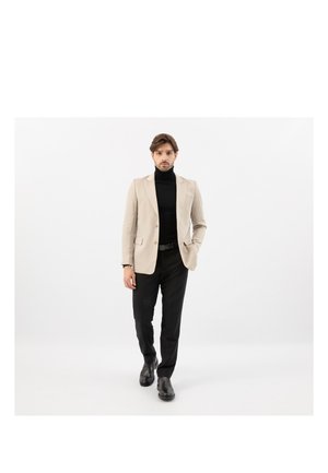 Uomo che cammina in avanti indossando un blazer beige, dolcevita nero, pantaloni neri e scarpe nere su uno sfondo bianco uniforme.