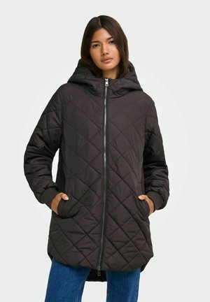Femme portant une veste matelassée noire à capuche avec fermeture éclair sur le devant et les mains dans les poches, associée à un jean bleu sur un fond uni.