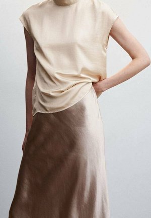 Kvinde iført en ærmeløs beige satin-top og en tofarvet brun satin-nederdel, stående med den ene hånd på hoften mod en ensfarvet baggrund.