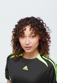 Nuori nainen, jolla on kiharat hiukset, pukeutuneena mustaan Adidas-paitaan, jossa on neonvihreät raidat, yksinkertaista vaaleaa taustaa vasten.