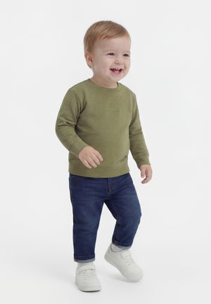Petit garçon souriant, portant un sweat-shirt vert olive, un jean bleu foncé et des baskets blanches, debout sur un fond blanc.