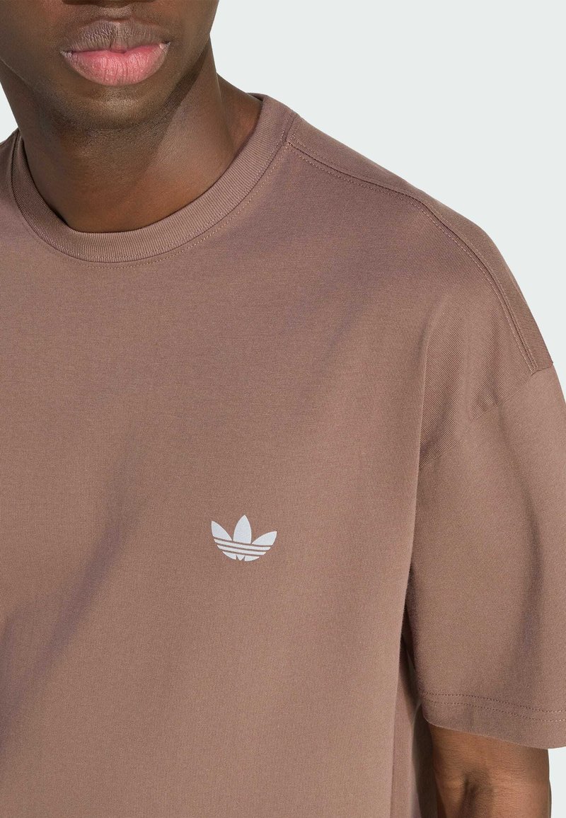 Homme portant un t-shirt marron discret à col rond avec un petit logo blanc Adidas trefoil sur la poitrine gauche.