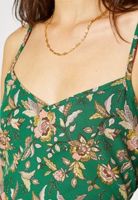 Top de tecido verde com padrão floral, possuindo alças ajustáveis, com flores em rosa, amarelo e branco, um decote em V e uma textura suave.