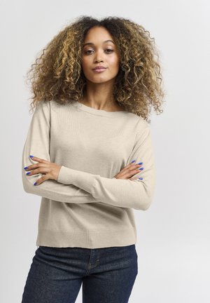 Pull en tricot beige avec un col rond, des poignets et un ourlet côtelés. Assorti à un jeans bleu foncé. Le modèle a des cheveux bouclés et des ongles bleus.