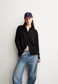 Schwarzes Langarm-Hemd mit Knöpfen, glattes Material, kombiniert mit hellblauen weiten Jeans. Marineblaues Cap mit weißen Logodetails.