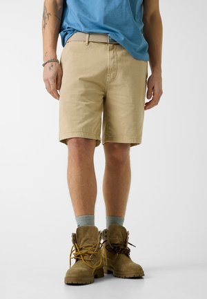 Man draagt beige korte broek, grijze sokken, bruine veterlaarzen, blauw overhemd gedeeltelijk ingestopt, staat tegen een effen achtergrond.