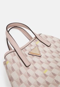 Sac à main beige et rose à motif avec deux anses roses et une plaque triangulaire dorée avec le logo Guess sur fond blanc.