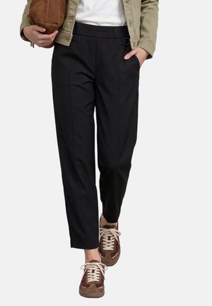 Personne portant un pantalon noir fuselé, des baskets marron et beige, une veste légère, et tenant un sac marron dans une main tandis que l’autre main est dans une poche.
