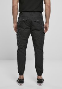 Pantalones cargo negros con puños elásticos, que presentan una tela texturizada, dos bolsillos traseros con cierres de botón y un diseño de pierna ajustada.