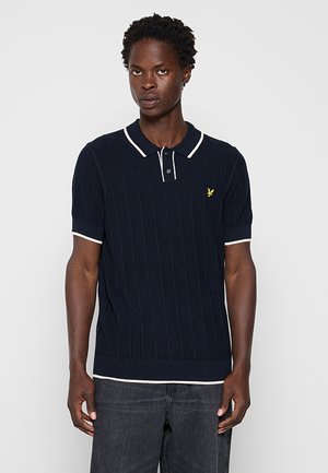 Mann trägt einen marineblauen kurzärmeligen Polo-Pullover mit weißem Besatz und einem kleinen gelben Vogel-Logo auf der Brust, kombiniert mit dunkler Jeans.