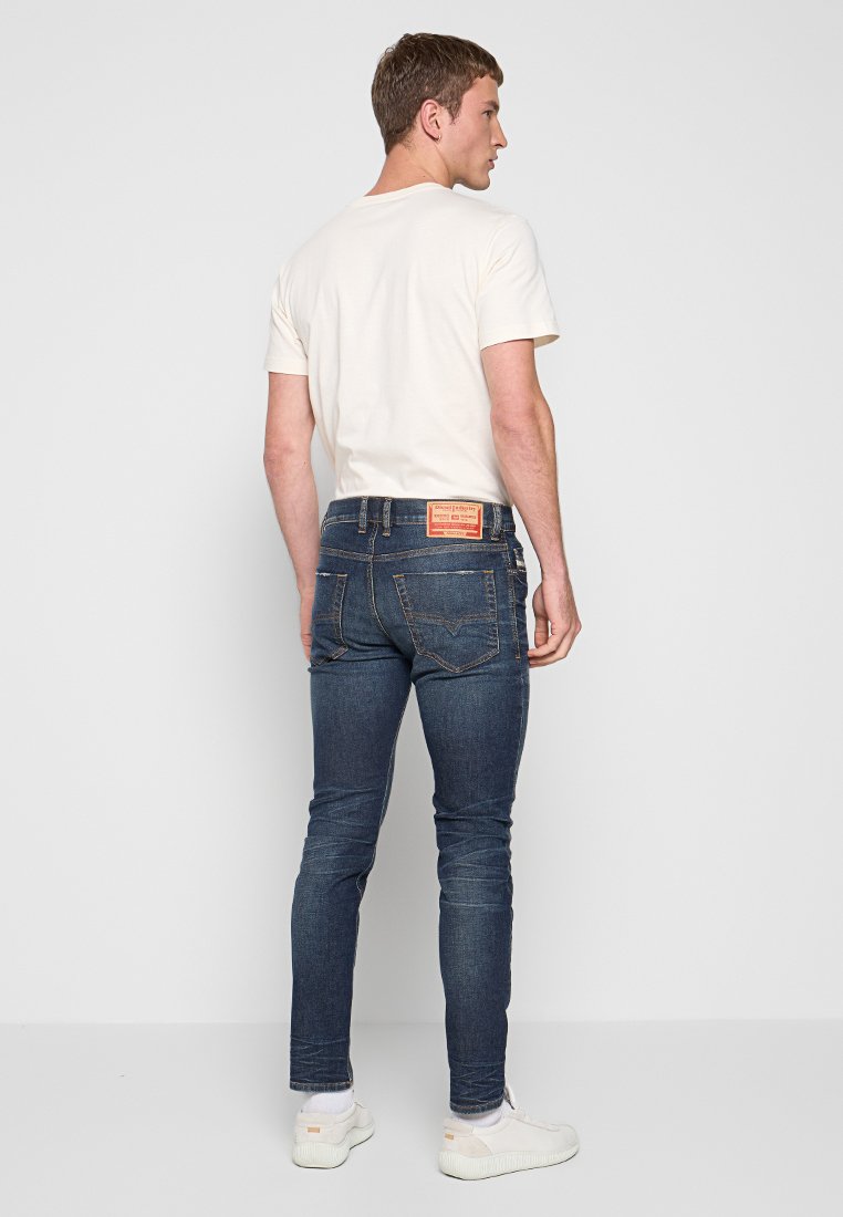 Mann trägt ein weißes T-Shirt, dunkelblaue Slim-Fit-Jeans mit einem Rückendesign, weiße Socken und weiße Sneaker, vor einem schlichten hellen Hintergrund.