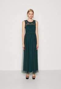 Rochie lungă de un verde închis, cu un corset din dantelă, design fără mâneci și fustă din tul fluidă. Asortată cu sandale cu platformă negre.